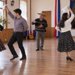 Dos parejas practican cueca mientras un instructor guía los pasos en un salón comunitario