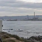 Equipo de monitoreo de calidad del aire frente a una bahía industrial en una imagen editorial referencial