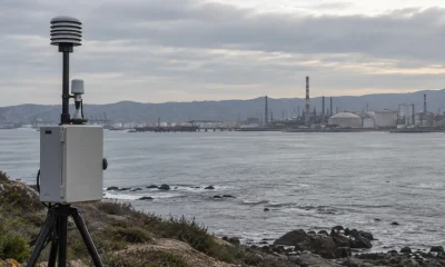 Equipo de monitoreo de calidad del aire frente a una bahía industrial en una imagen editorial referencial