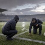 Trabajadores revisan una cancha de fútbol bajo la lluvia antes del inicio de la fecha 16