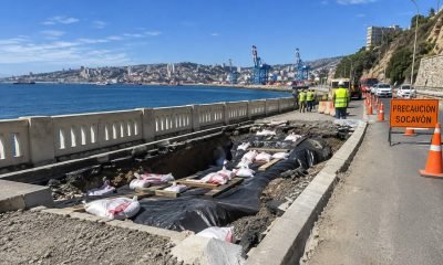 Socavón junto a una vía costera, con obras, conos y una señal de precaución.