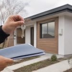 Manos con llaves y documentos frente a una vivienda nueva