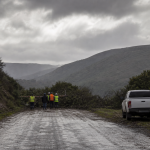 Camino rural húmedo con personal de emergencia visto a distancia tras un sistema frontal, imagen editorial referencial